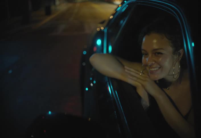This lowlight video features a woman leaning out of a car window at night on a city streetshe smiles and laughs clearly enjoying herself as the streetlights and bokeh from traffic illuminate her face