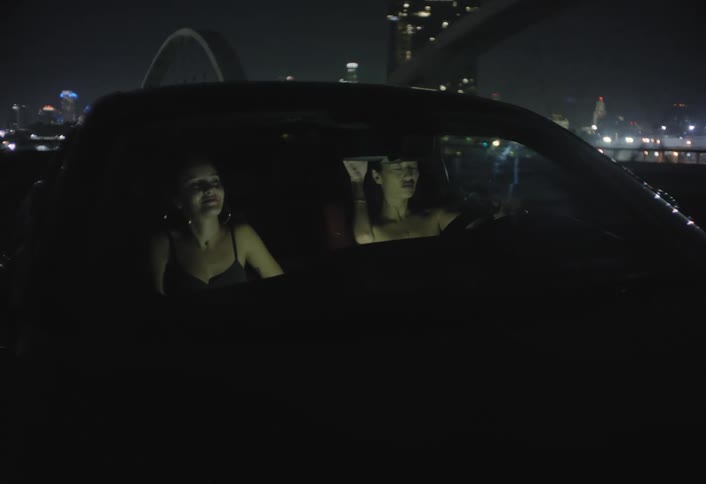 This clip features a front exterior shot of a car at night showing two women in the front seatsthey are smiling laughing and gesturing as they enjoy the ride with the glowing city skyline visible in the background