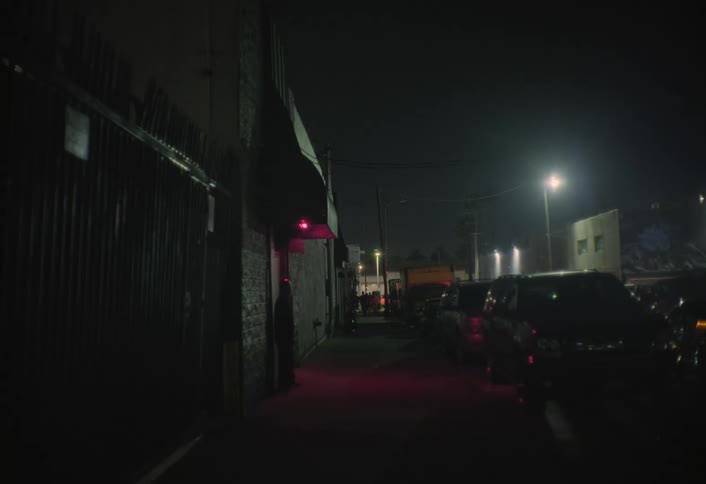 A city street at night with several parked cars and a lamppost with a white light in the background