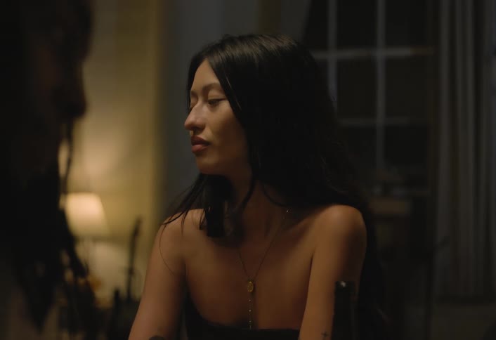 A woman with long dark hair seen in profile turns her head while sitting and talking in a dimly lit room at night
