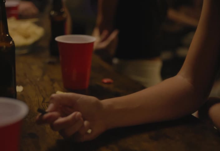 A closeup shot pans from a womans face down to her hand as she removes the cap from a vape pen on a table with red cups