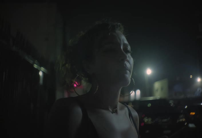 A young woman in a dark top is standing on a dimly lit street at night as she holds a phone before bringing a cigarette to her mouth and exhaling with blurry city lights providing highcontrast backlighting in the background