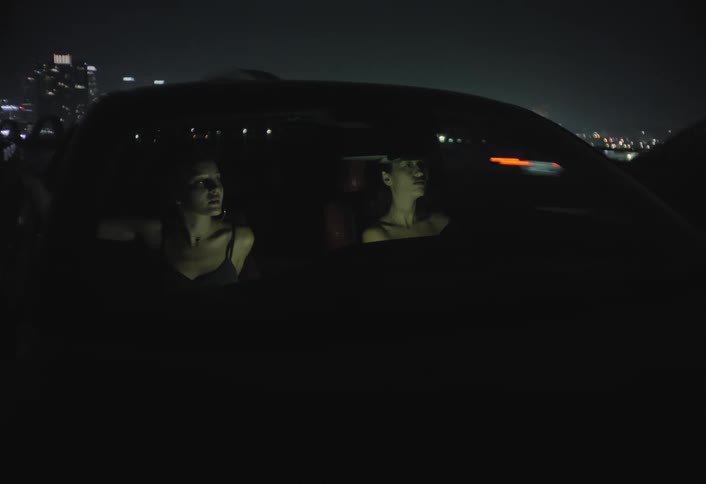 Two young women a driver and a front passenger are seen from a vantage point outside the front windshield of a vehicle at night driving through a city landscape characterized by distant lights and infrastructure