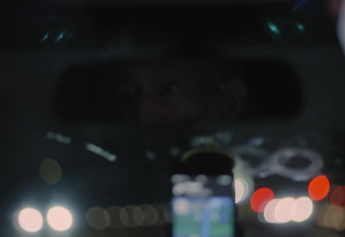 This short dark clip is shot from the drivers perspective inside a car at nightthe foreground is blurred by bokeh from streetlights and the cars rearview mirror with a cell phone displaying a map app mounted below it