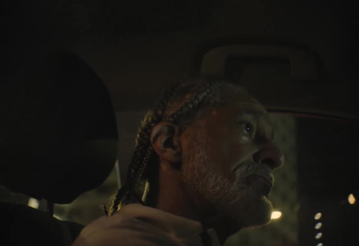 This is a dark lowangle shot of a man with braided hair and a prominent lightcolored beard sitting inside a car at night streetlights and illuminated building windows provide subtle rim lighting that catches the side of his face