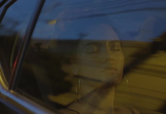 A woman naps in the back seat of a moving car at night her face visible through the window as city lights reflect and pass by