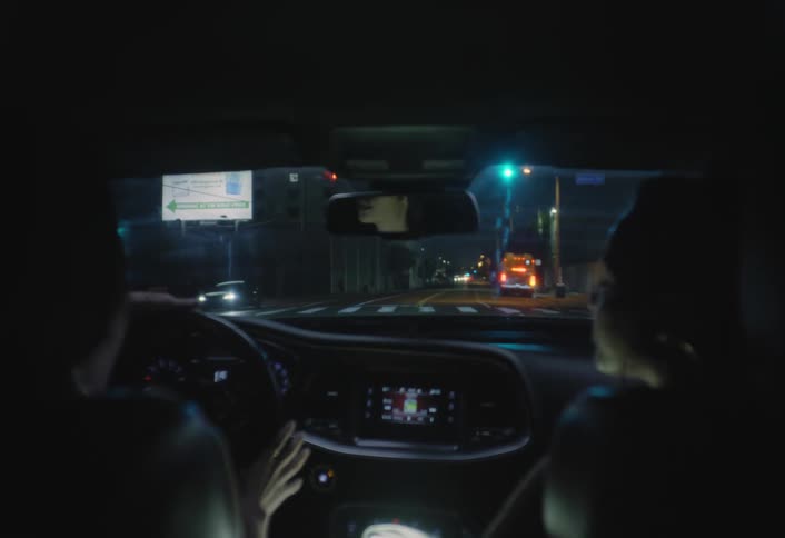 The video shows a dark nighttime view from the back seat of a car as it drives through a city traffic lights street lamps and the headlights and taillights of other vehicles illuminate the road ahead