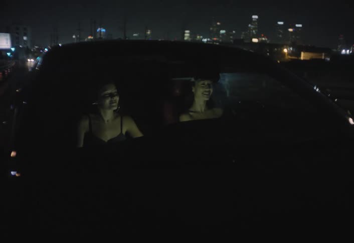 Two young women seated in the front seats of a vehicle at night are seen through the windshield exchanging smiles and looking at each other while the car is moving slowly through city traffic