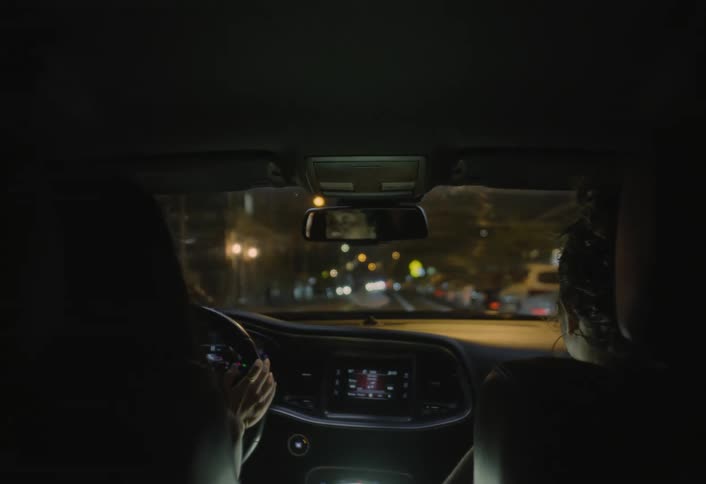 A car interior at night shows a driver and passenger in silhouette as the dashboard glows and distant city lights shimmer through the windshield