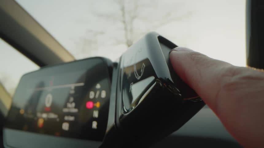 A closeup shot focuses on a finger pressing the button on a gear selector stalk inside a modern car likely an electric vehicle during a bright day in the background the digital instrument cluster shows the current vehicle status and driving mode