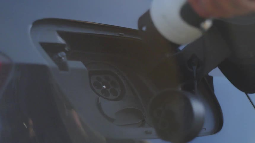 A closeup shot shows a hand disconnecting a yellowandblack charging cable from the charge port of a blue electric vehicle the port cover remains open revealing the circular charging contacts inside