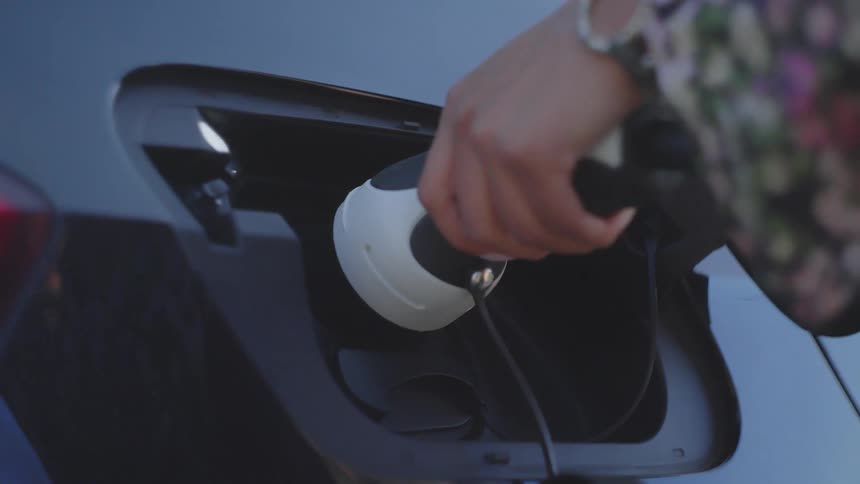 A closeup shot shows a hand inserting a charging connector into the open charge port of a dark electric vehicle successfully securing the plug the shot features the cars blue exterior and the persons patterned sleeve and wrist accessories
