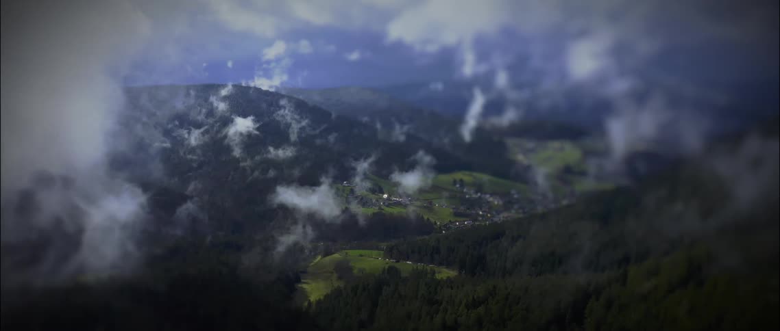 An aerial shot captures a mountain valley vista as wisps of mist and clouds slowly move across the frame partially revealing and concealing the landscape below a small village and patches of green pasture are nestled among the dark dense forest coveri