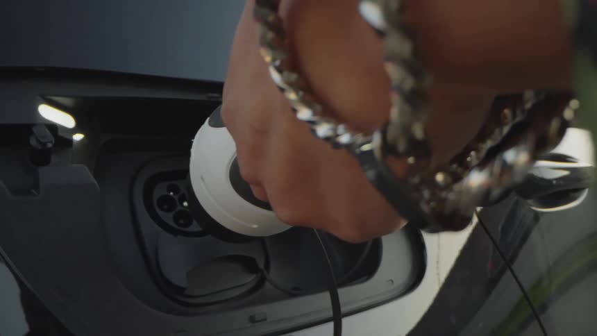 A closeup shot focuses on a persons hand plugging a white and black charging connector into the open charge port on the side of a darkcolored electric vehicle the hand is wearing a bracelet and carefully guides the plug into the socket