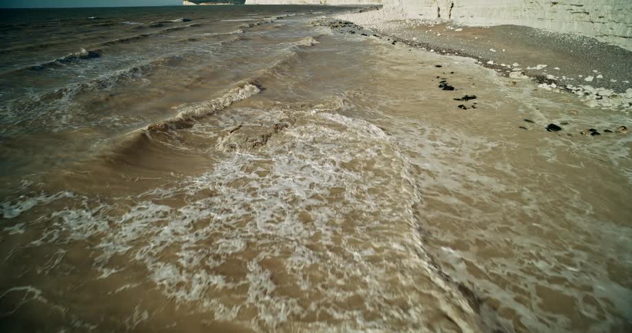An aerial shot shows brown ocean water washing up onto a beach of gray stones at the base of dramatic white chalk cliffs