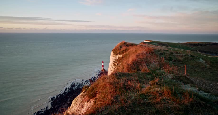 A lighthouse stands at the base of a sunlit grassy cliff overlooking the ocean