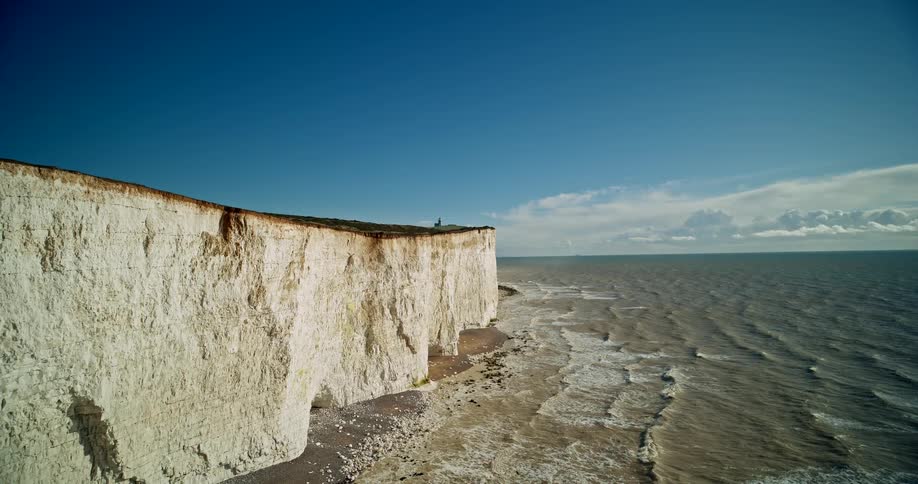 An aerial view captures the dramatic white chalk cliffs towering over a rocky beach and the deep blue ocean