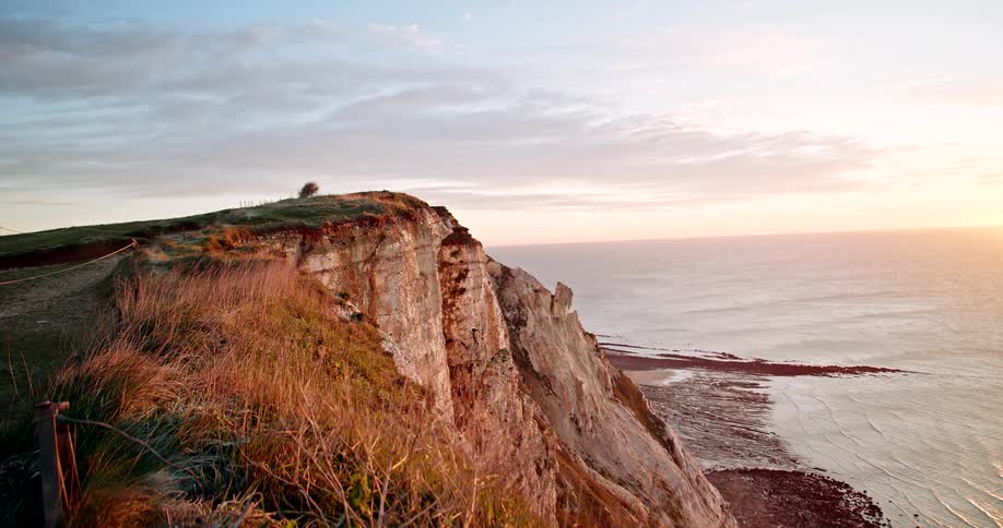 A cliff stands tall above a beach as ocean waves wash upon it at sunset