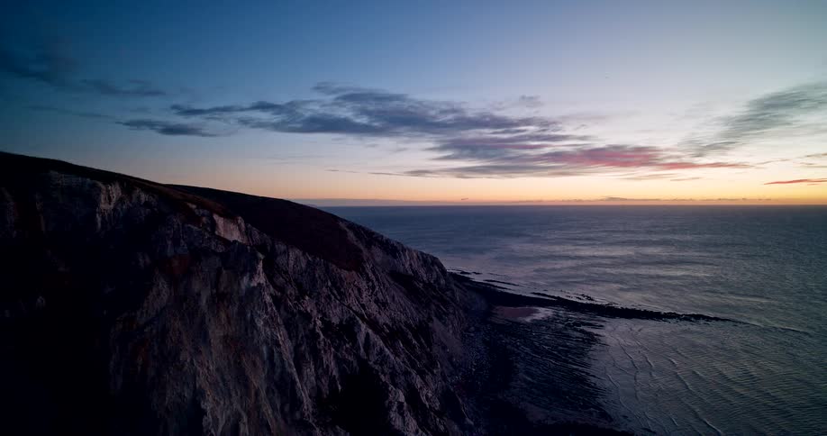 A dramatic aerial shot captures the steep rugged slopes of a dark cliff plunging into the ocean with the horizon lit by the vivid colors of a dawn or dusk sky