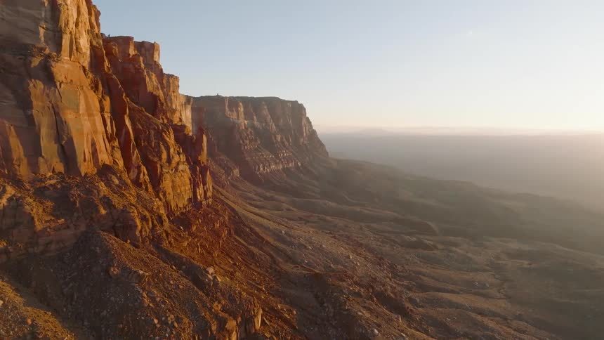 An aerial shot sweeps past a towering sheer red sandstone cliff face and a dramatically sloping arid landscape the warm lateday sunlight intensely illuminates the rock formations casting a golden glow over the desert canyon environment