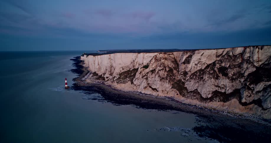 An aerial shot shows a stretch of pale chalk sea cliffs with striped lighthouse at blue hour