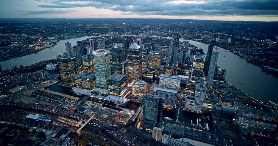 A high aerial shot captures a dense cluster of illuminated skyscrapers in a modern financial district surrounded by a wide river at dusk