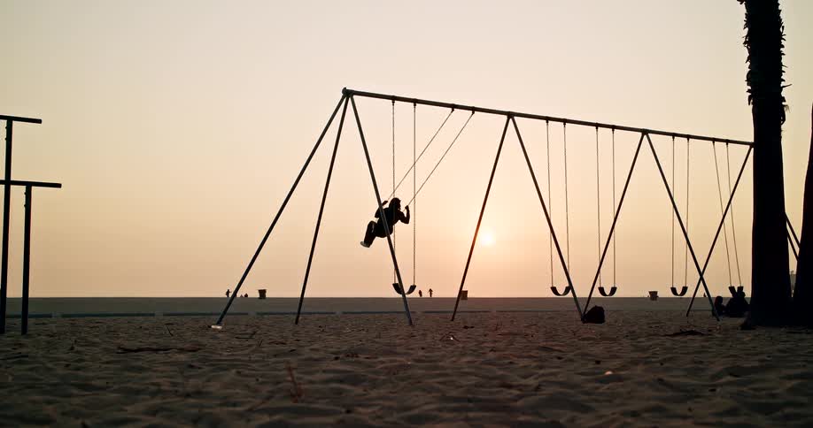 The video shows a silhouette of a person swinging high on a swing set located on a sandy beach at sunset the low sun creates a warm orange and yellow haze over the flat horizon with the swing set and a nearby palm trunk framed in dark shadow