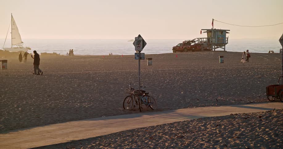People walk on a beach and ride a bike as a sailboat goes past in the water