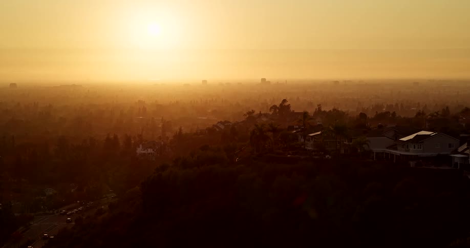An aerial shot captures a vast sprawling city landscape viewed from above a forested hillside neighborhood during a vibrant sunset or sunrise the entire scene is bathed in a thick goldenorange haze with a distant skyline faintly visible on the horizon