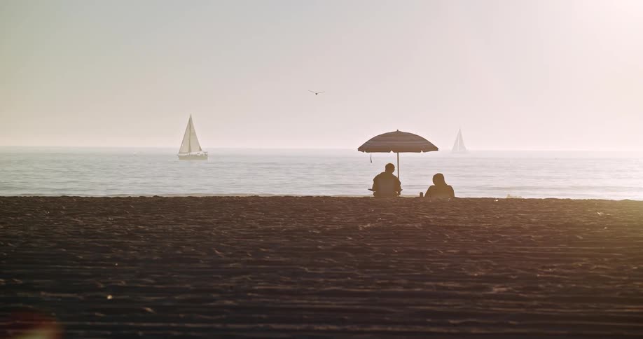 The silhouette of two people under a parasol on a beach one morning looking out to sea with some sailboats