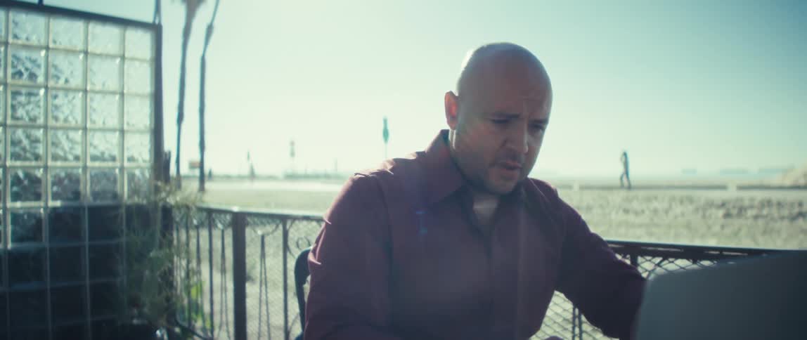 A man sits with his laptop on an outdoor patio near the beach