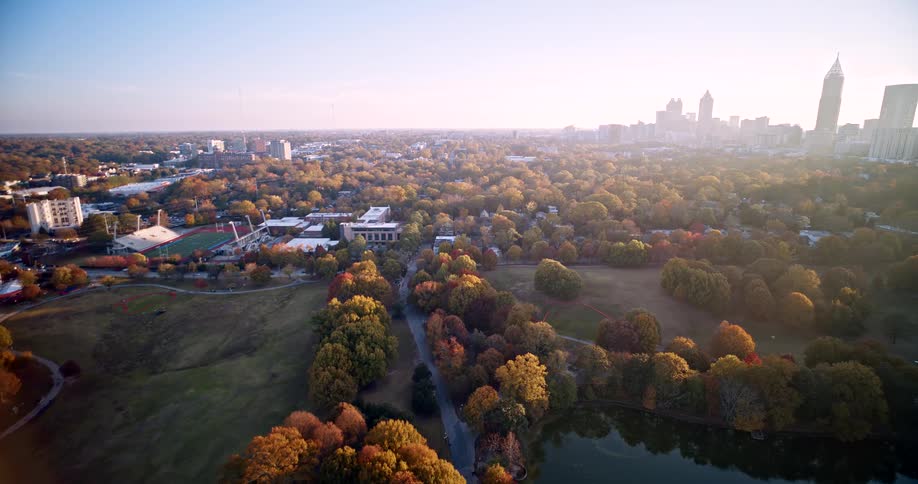 This aerial shot provides a sweeping view of a large urban park and a university campus with the cityscape fading into the distance on the right the foreground is dominated by trees showcasing vibrant fall foliage and a small body of water suggesting a