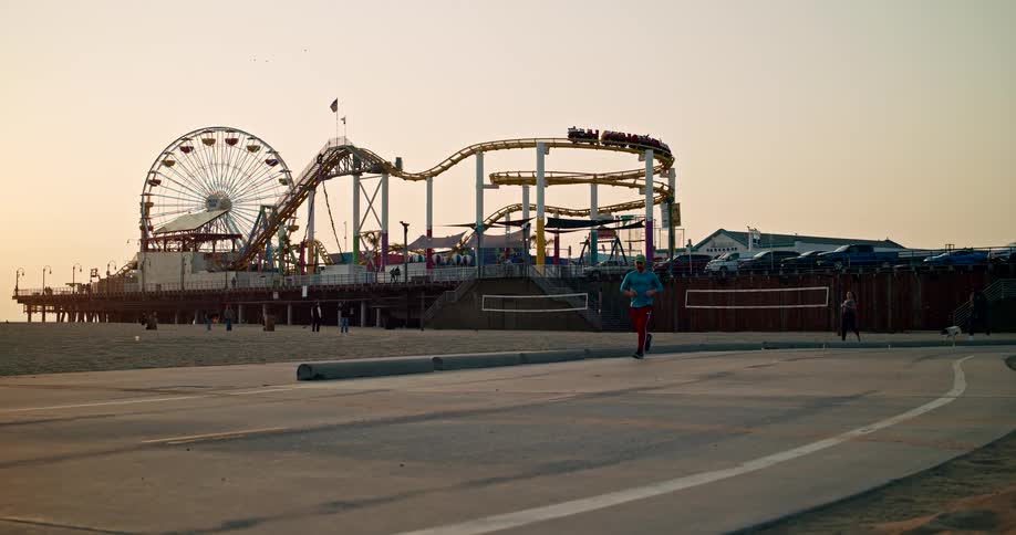 People jogging on a road outside of an amusement park as a rollercoaster filled with riders rolls around a curve