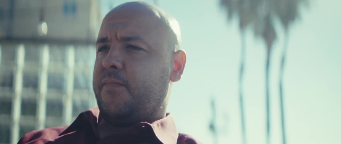 The video shows a closeup slightly lowangle shot of a bald man with a beard wearing a reddishbrown collared shirt he is outdoors on a bright sunny day with the sunlight creating a strong contrast and slightly overexposed look especially highlight