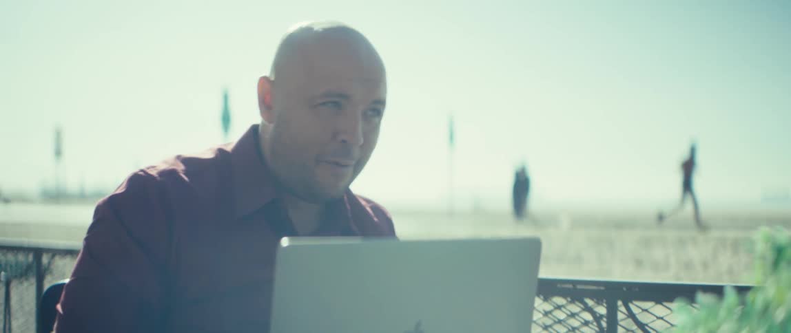 A bald man smiles while using a laptop outdoors with a bright hazy beach in the background