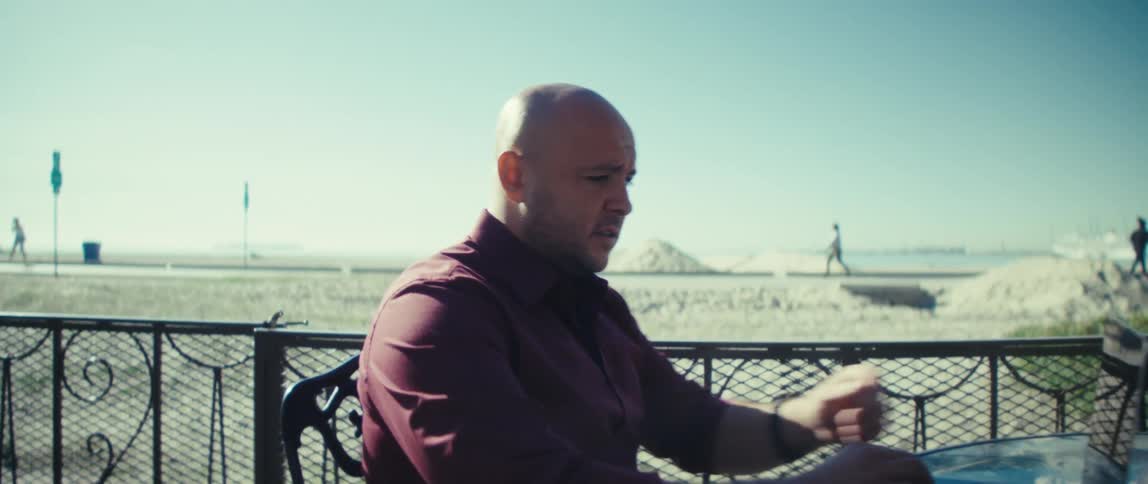 Man sitting at a table on a balcony typing on a laptop computer as he stops to look at his phone