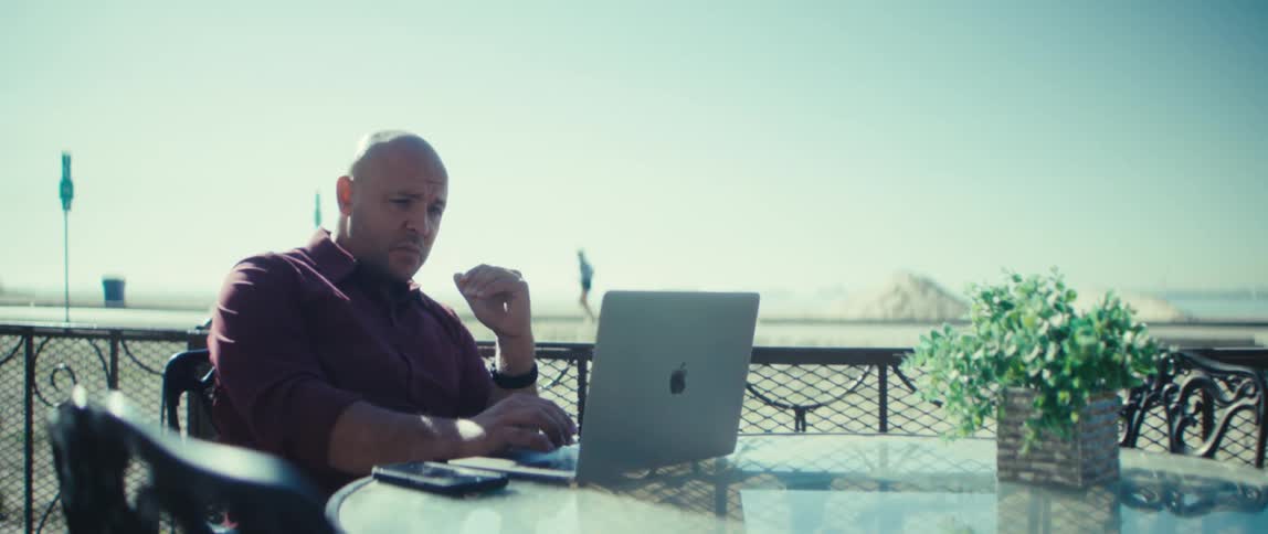 A bald man is sitting at an outside table talking in front of a laptop