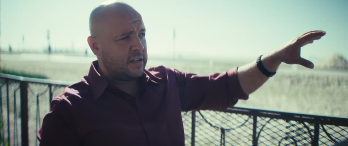 A bald man sits outdoors with a laptop speaking and gesturing passionately