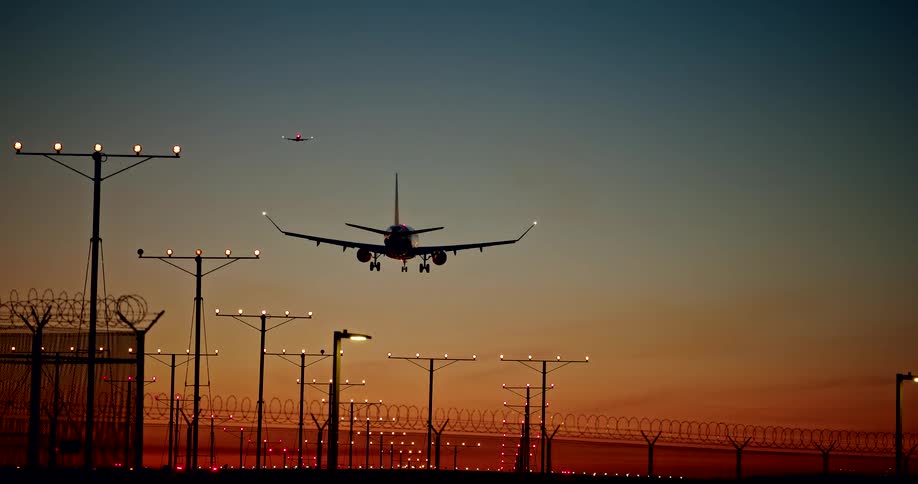 Silhouetted against a sunset sky an airplane descends for landing over an illuminated runway