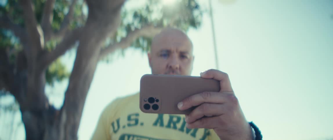 This video uses an extreme lowangle shot looking up at the same bald man who is holding a modern smartphone in a brown case horizontally seemingly recording a video or taking a photo he is wearing a yellow shirt with us army vet visible on the che