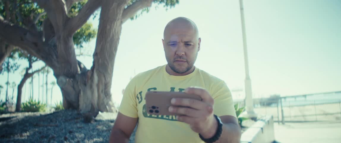 A man is sitting outside under a tree as he looks at his cellphone