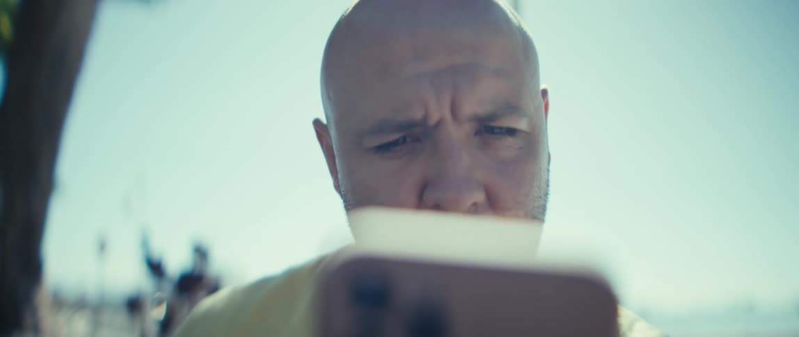 A very closeup shot focuses on the face of a bald man looking down intensely at a phone he holds below the frame causing him to furrow his brow in concentration the bright sunlight creates a highkey visual with a strong washedout background