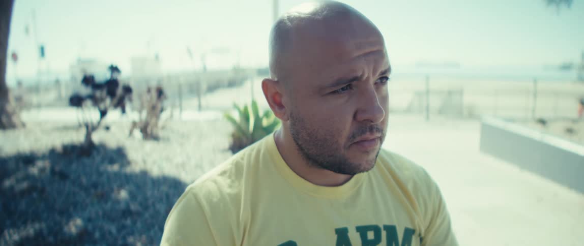 A man is sitting near a beach where people are on the boardwalk