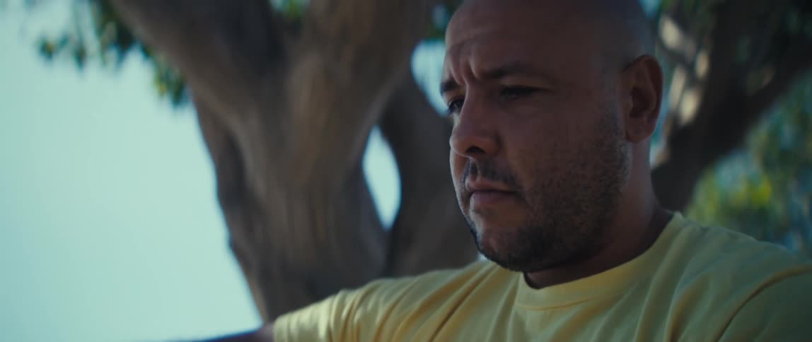 A bald man with a short beard wearing a yellow shirt with the word army on it sits outdoors under a tree looking intently at the screen of a mobile phone he holds in his hand the bright sunny light catches the side of his face as he appears to be r