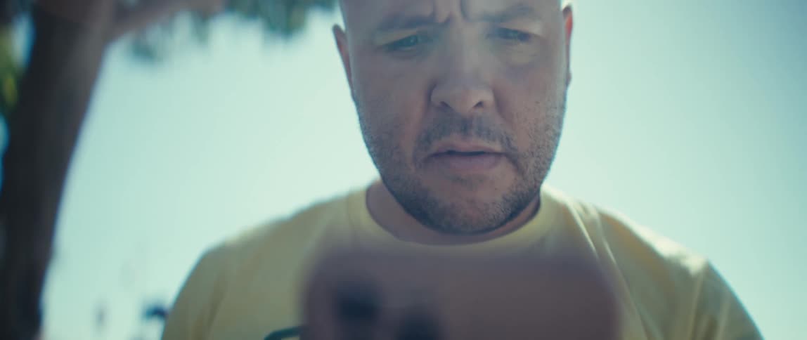 A young man wearing a yellow tshirt looks at his mobile device under the midday sun