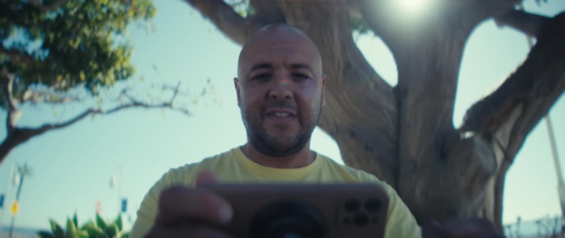 Young bald man wearing a yellow shirt smiling and cheering while watching something on a smartphone on his hands outdoors during day time