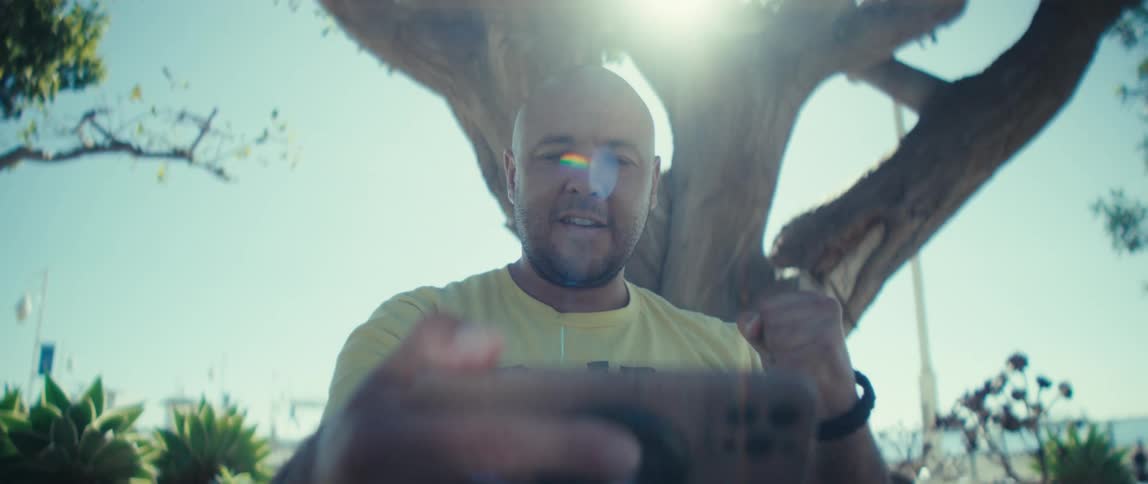Bald young man wearing a white shirt smiling and cheering while looking at a smartphone on his hands outdoors during day time