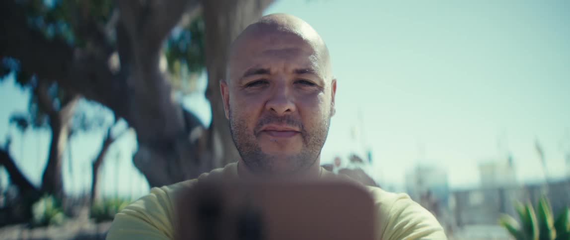 Shavedhead person in a yellow shirt holds a smartphone in a treelined outdoor area