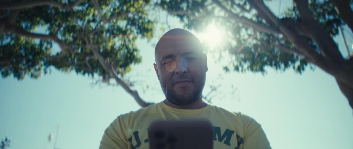 A man is wearing a yellow us army veteran shirt holds a phone in front of him looking down at the screen for most of the clip the camera is angled up towards him and the sky capturing bright sunlight that shines through the leaves and bran