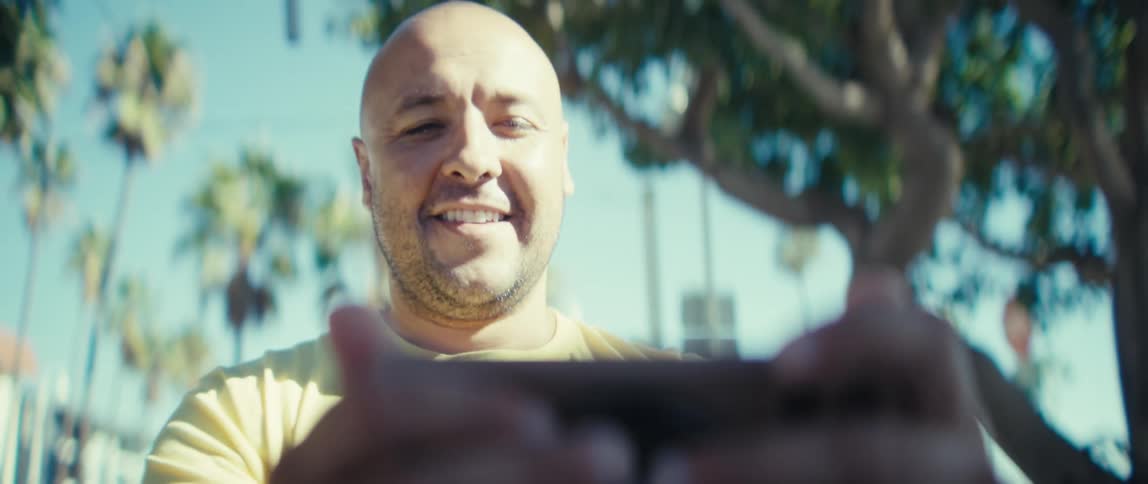 Man in a yellow shirt watches content on a smartphone with blue sky and palm trees in the background